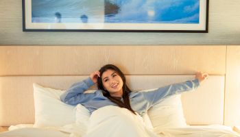A woman relaxing on a bed in a hotel room, leaning back with pillows, wearing a denim shirt and white sheets around her, smiling.