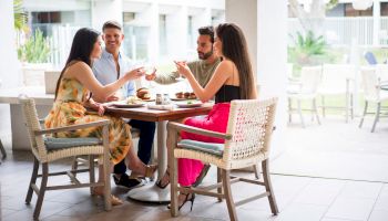 A group of four friends sits around a table on a sunny patio, chatting and toasting with drinks and plates of food.