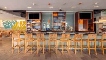 A bright, modern bar with a long wooden counter and row of high chairs, shelves of drinks behind, and a coastal, airy vibe.