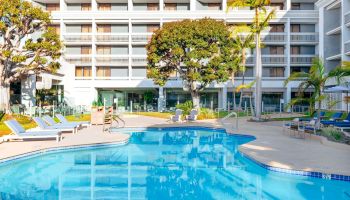 A bright hotel pool area with a large blue swimming pool, lounge chairs, palm trees, and a multi-story white building in the background.