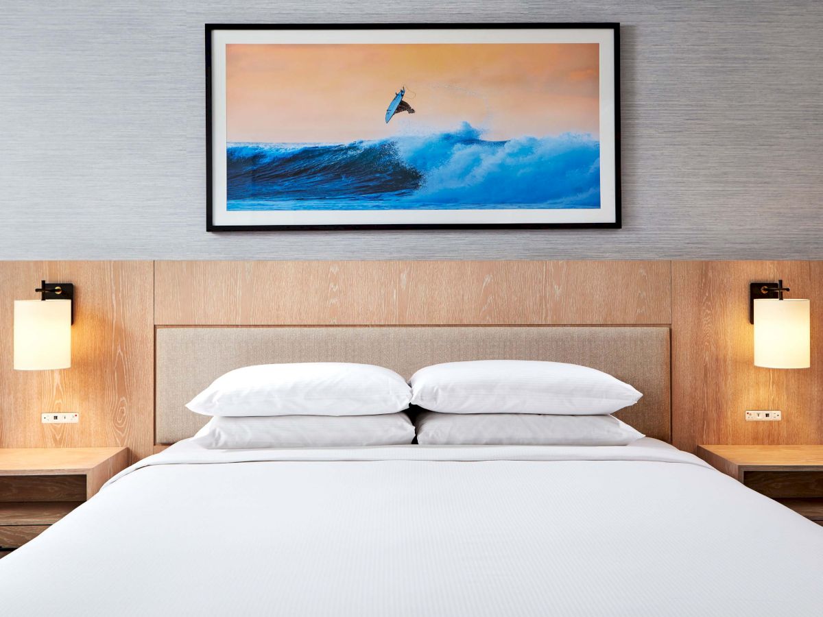 A neatly made hotel bed with white linens, nightstands, wall lamps, and a framed ocean photo above the headboard.