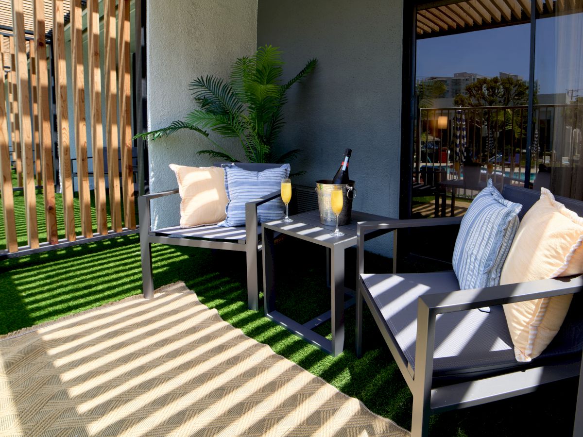 A small outdoor seating area with two dark chairs, beige cushions, a matching table, drinks on the table, and a potted plant, under a shaded patio.