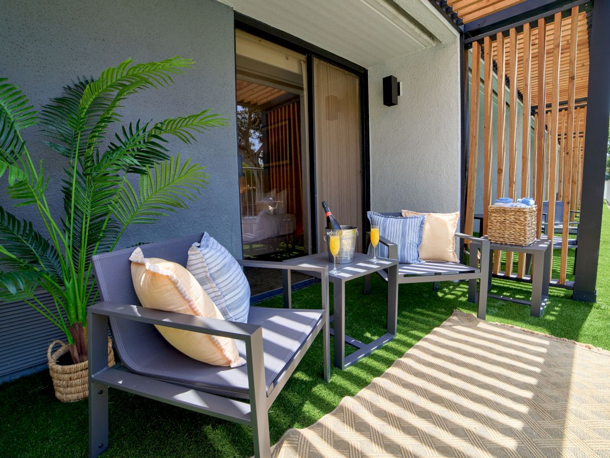 A cozy outdoor seating area with two cushioned chairs, a small table, potted plants, and a shaded patio under a wooden pergola.