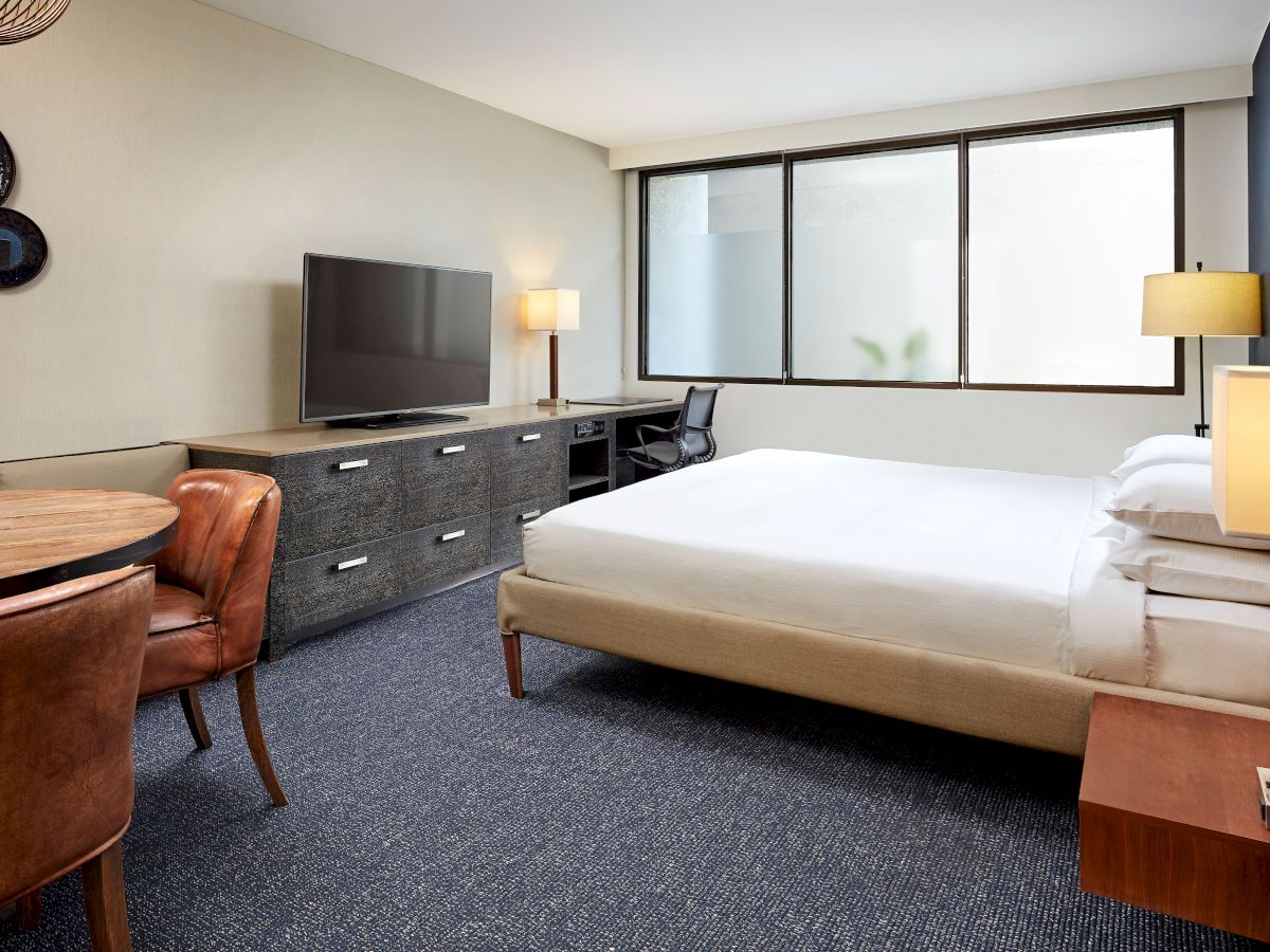 A modern hotel room with a king bed, a desk, a flat-screen TV, dresser, two lamps, and a windowed wall; warm wood accents and blue carpet.