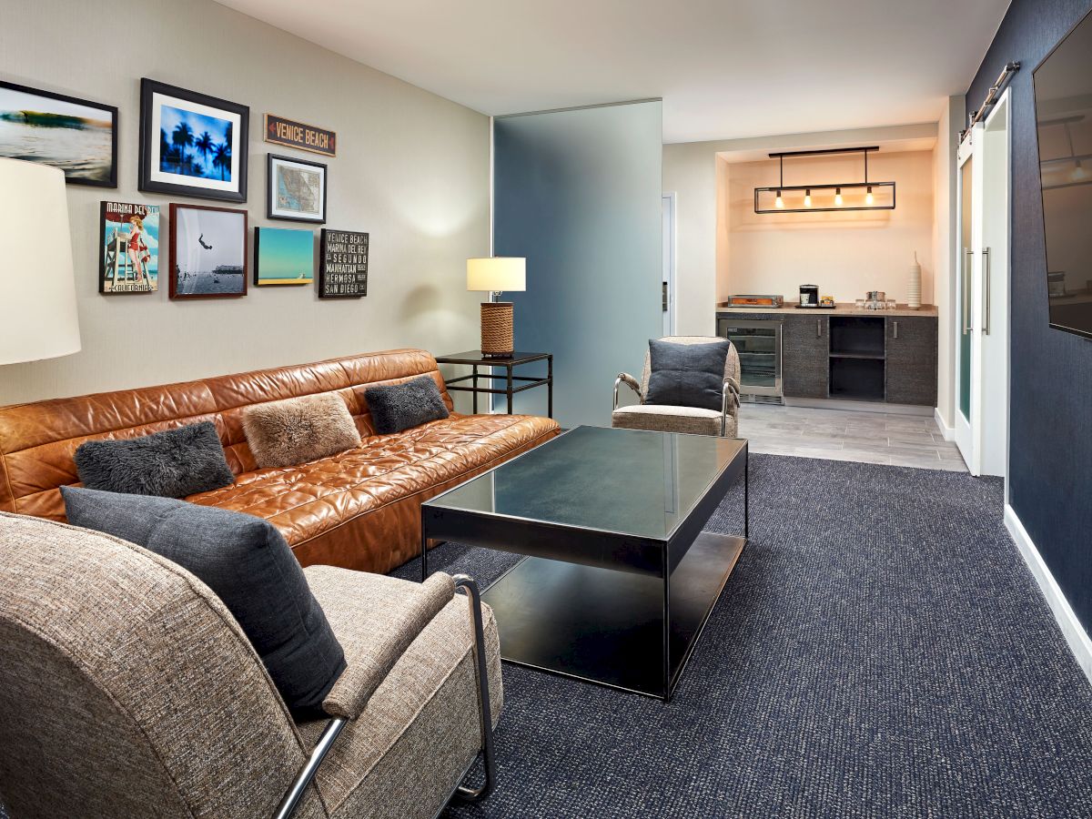 A cozy living room with a tan leather sofa, gray armchairs, a coffee table, framed wall art, and a hallway leading to a kitchenette.