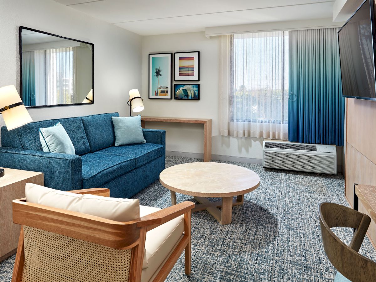 A cozy hotel living room with a blue sofa, armchairs, round coffee table, wall art, a desk, big window, and a wall-mounted TV, neat and inviting.