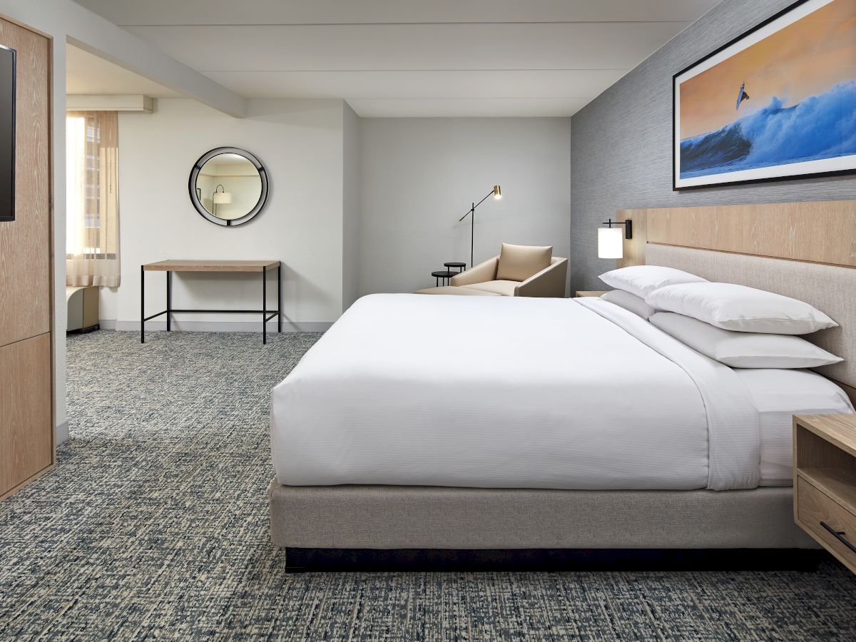 A modern hotel room with a large bed, nightstands, a bench and desk, wall art of a surfer, a round mirror, and a cozy seating area in the back.