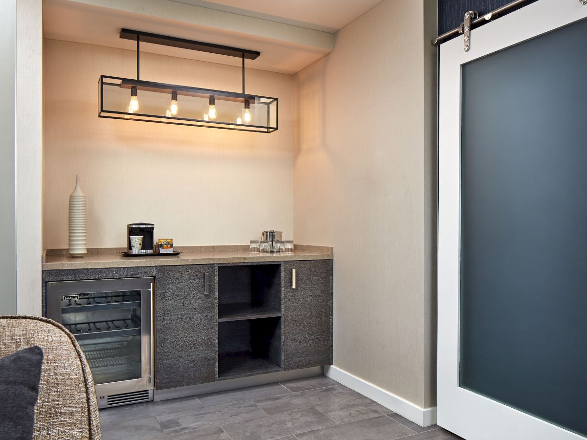 A small kitchenette with a counter, mini fridge, open shelving, a sink, and a modern pendant light&mdash;next to a frosted sliding glass door.