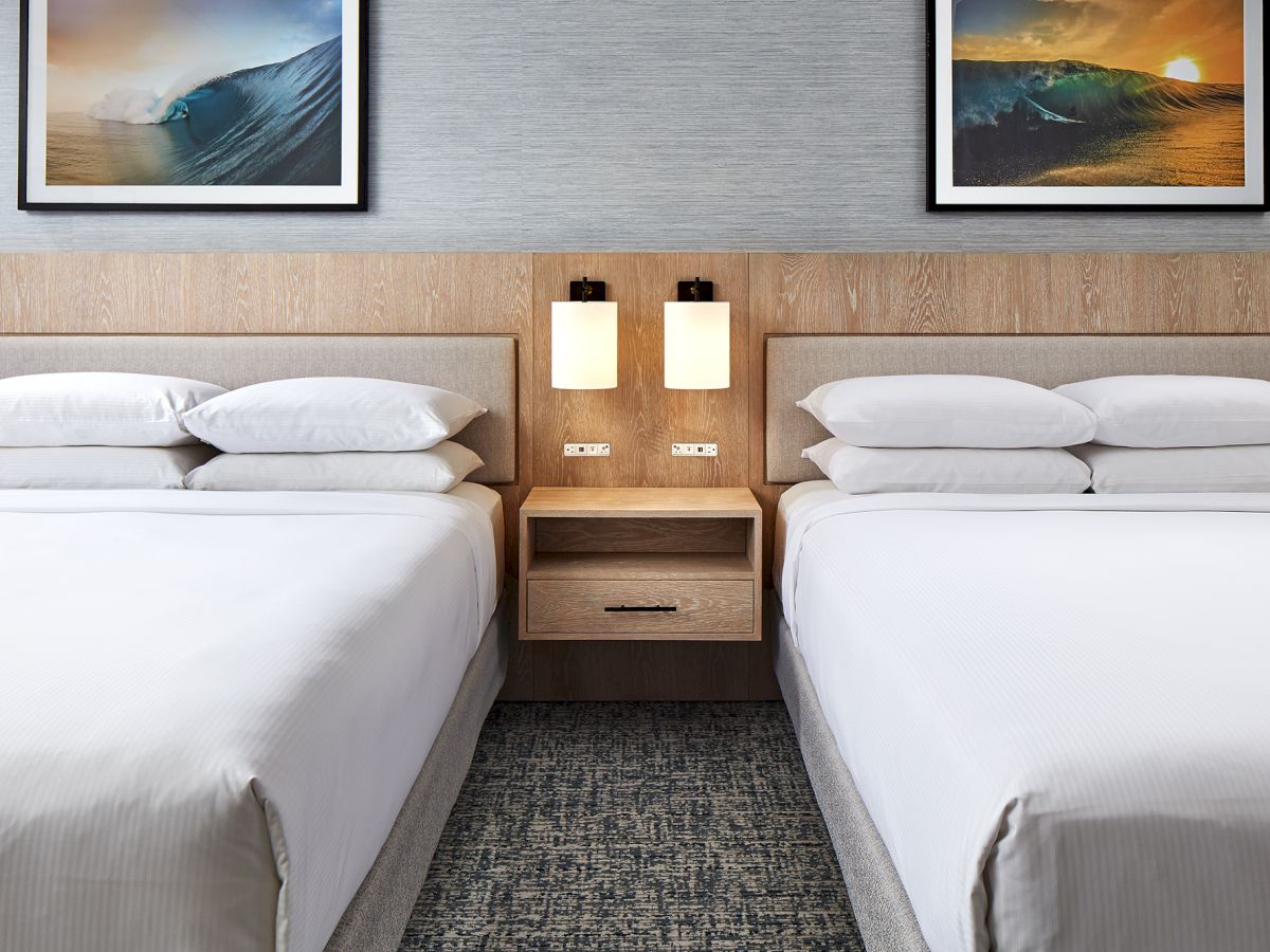 Two neatly made hotel beds with a shared nightstand and lamps between them, framed art above showing ocean waves.