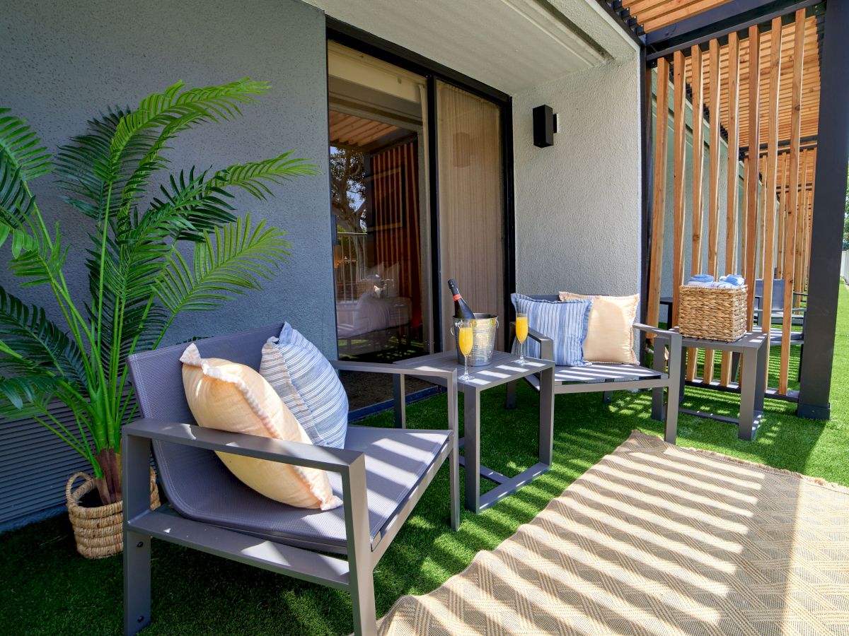 A cozy outdoor seating area with two chairs, cushions, a small table with drinks, a potted plant, and a shaded patio beside a house.