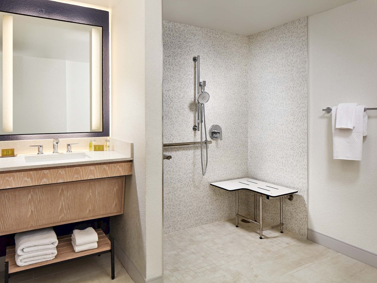 A modern bathroom with a wooden vanity, lit mirror, neatly stacked towels, and a spacious walk-in shower featuring a bench and handheld showerhead, plus a wall-mounted towel bar.