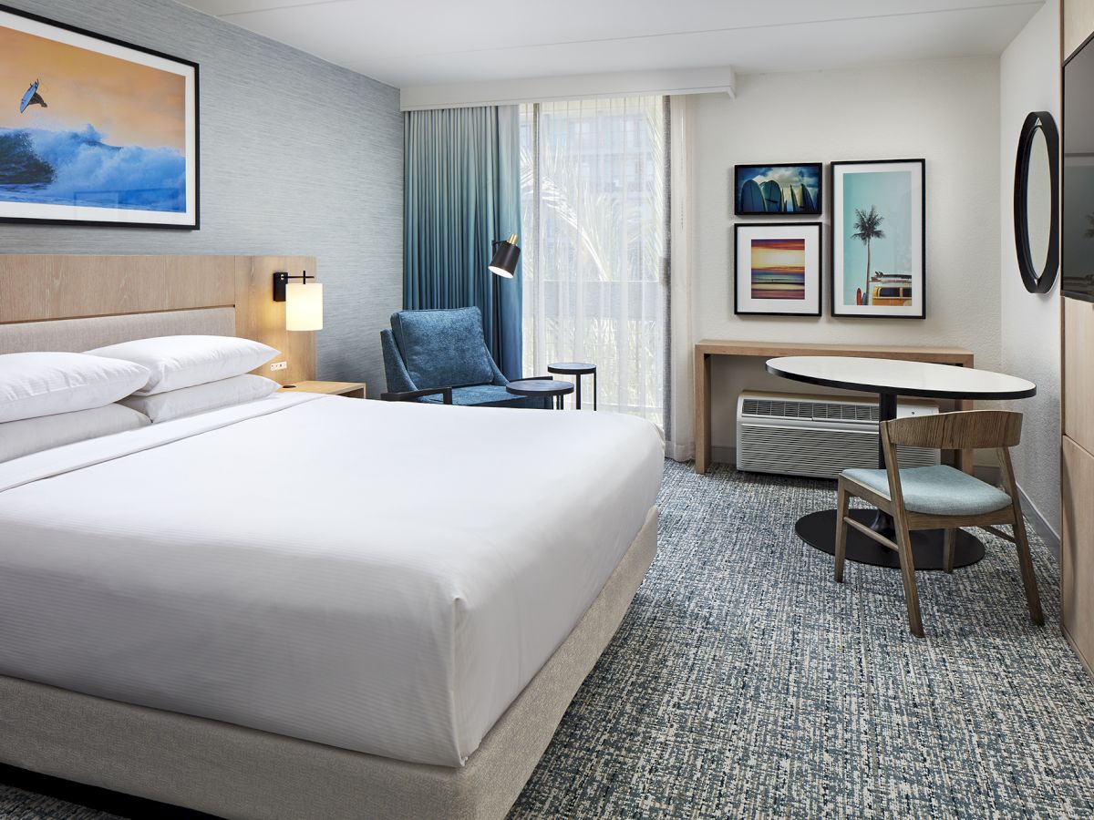 A modern hotel room with a large bed, blue chair, wall art, desk, round mirror, and a window with curtains, all in a neutral palette.