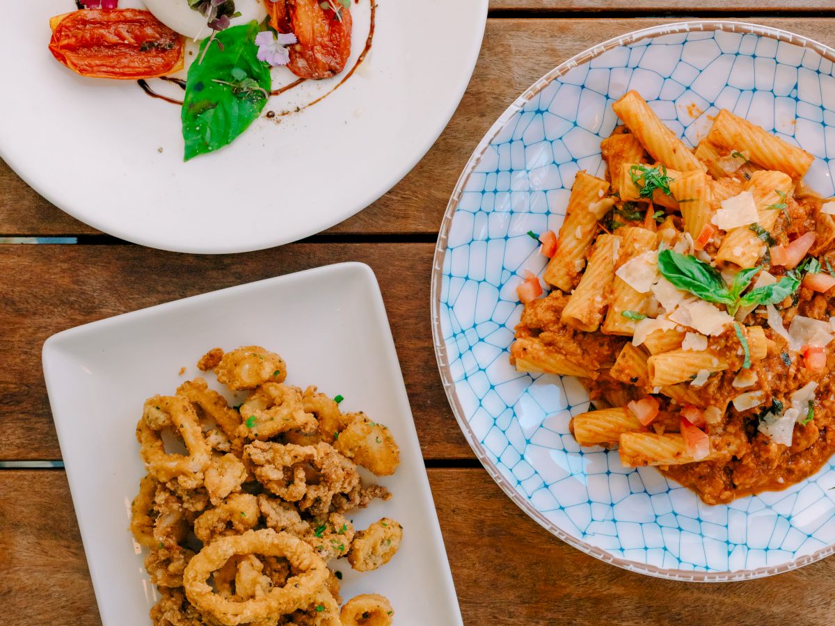 A trio of gourmet plates: crispy fried calamari with dipping sauces, pasta in a rich tomato sauce, and a fresh herb salad over grilled peaches.