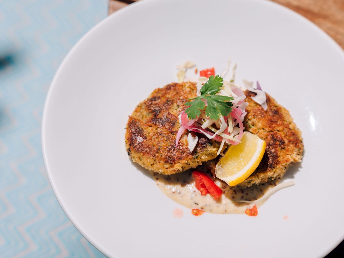 Two golden-brown croquettes on a white plate, topped with red onion, cilantro, and small peppers, with a lemon wedge and creamy sauce beside.