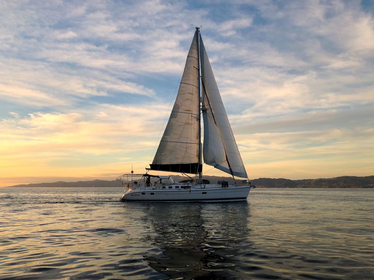 A sailboat glides over calm water at sunset, its sails full as a gentle breeze paints the sky with warm hues.