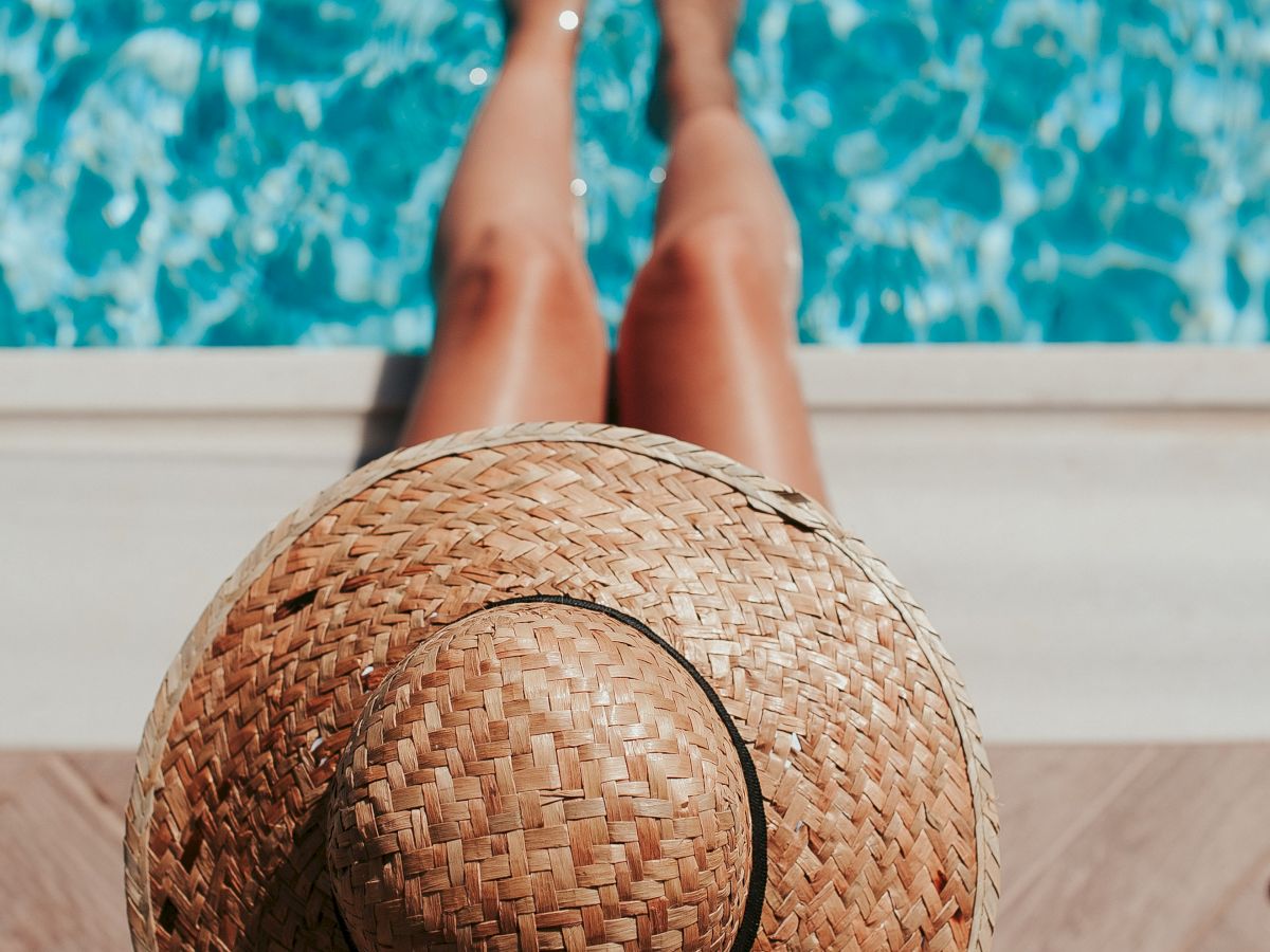A person wearing a hat sits at the edge of a pool with their legs dangling over the water, sunny vacation vibes, ending with a period.