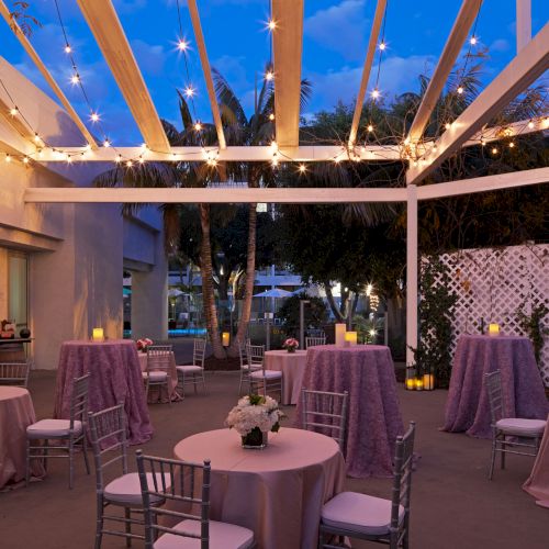 A quaint outdoor dining courtyard with string lights, wooden tables, and chairs, set against a warm brick building and evening sky.