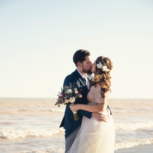 A couple on a beach kiss, bride in a white gown holding a bouquet, sunset-lit waves in the background, romantic and joyful moment.
