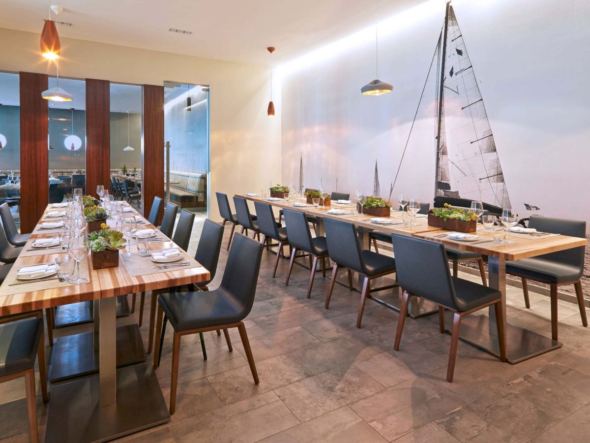 A modern dining room with long wooden tables, navy chairs, small potted plants, pendant lights, and a sailboat mural on the wall.