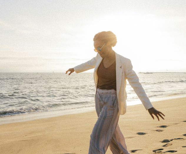 A person walks along a sunny beach at the shore, toes in the sand, posing with arms outstretched as the ocean glistens nearby.
