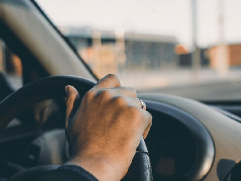 A close-up of hands gripping a car steering wheel from the driver&rsquo;s seat, with a blurred road scene outside the windshield.
