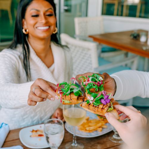 Two friends share a colorful, decorated burger feast outdoors; one hands over a vibrant, green-pasted mini burger to the other, smiles all around.