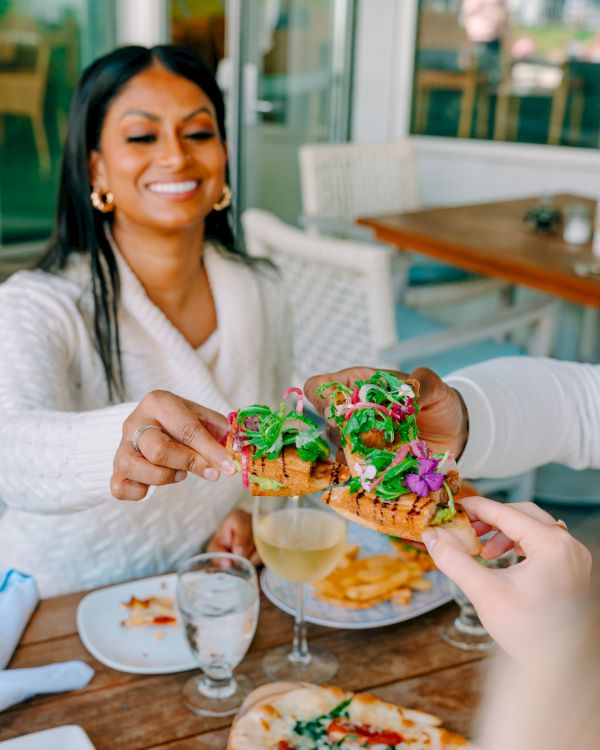 Two friends share a colorful, decorated burger feast outdoors; one hands over a vibrant, green-pasted mini burger to the other, smiles all around.