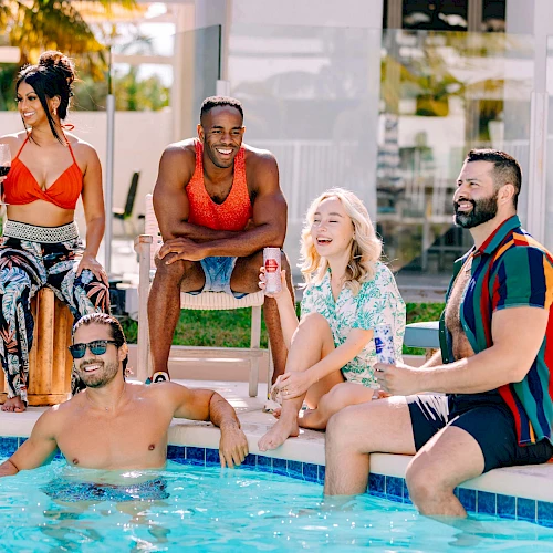 A group of stylish friends relax by a pool: some sit on the edge, one in the water, others stand or chat, enjoying a sunny day together.