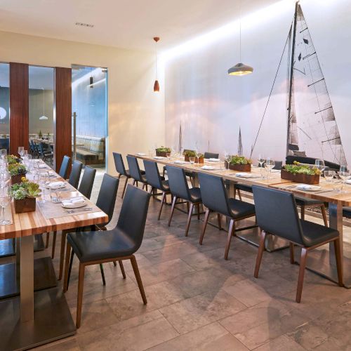 A modern dining room with long wooden tables, blue chairs, small plants, pendant lights, and a nautical wall mural featuring a sailboat.