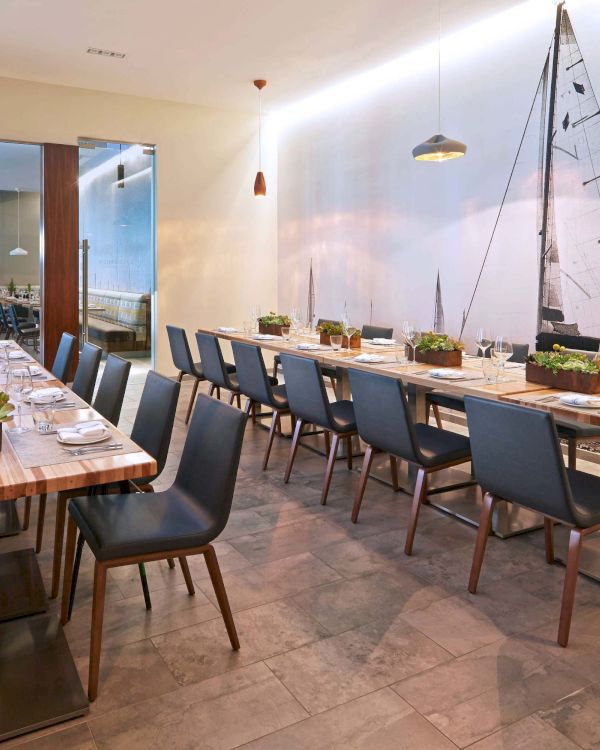 A modern dining room with long wooden tables, blue chairs, small plants, pendant lights, and a nautical wall mural featuring a sailboat.